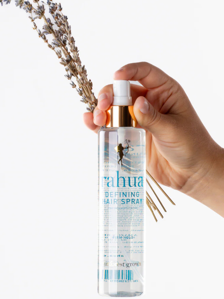 A hand holding a clear bottle of Rahua Defining Hair Spray with a gold spray top, and a small bouquet of dried lavender is visible in the background.