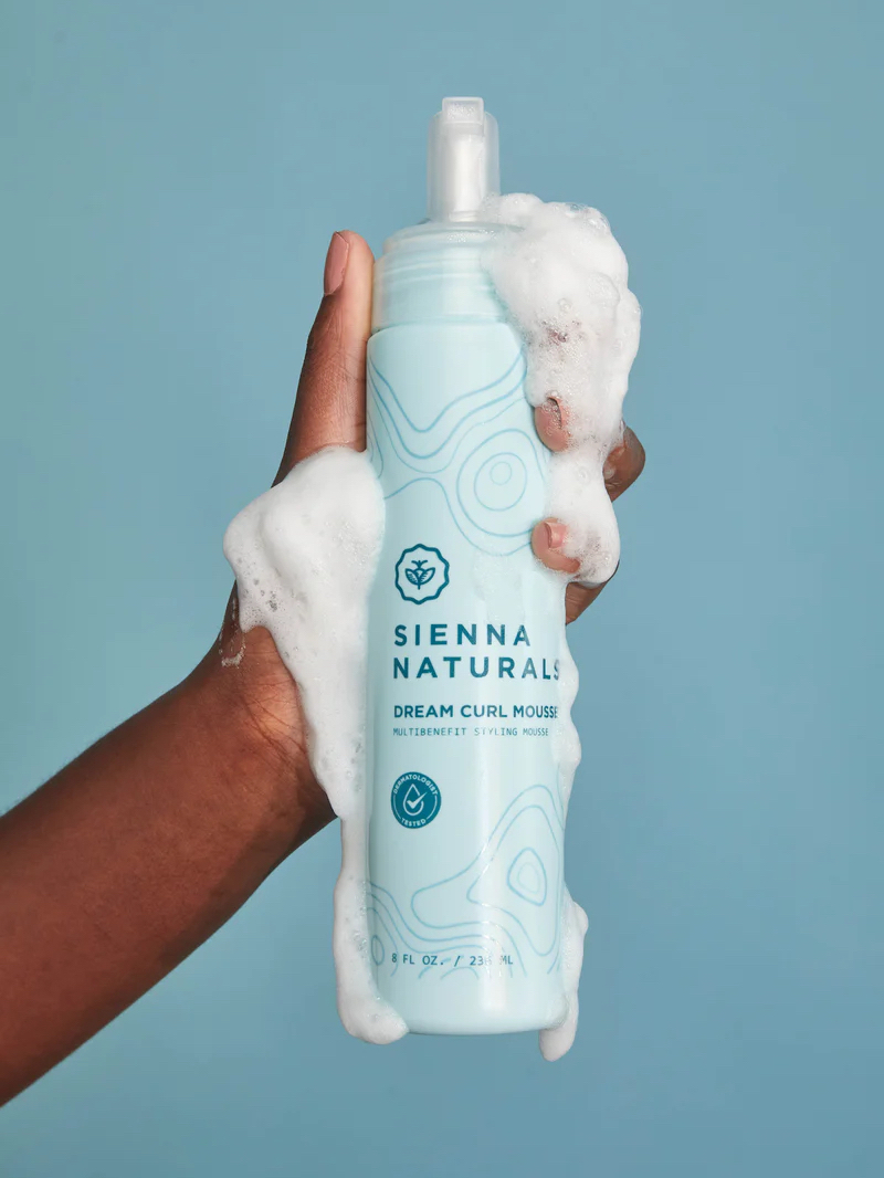 A hand holding a blue bottle of Sienna Naturals Dream Curl Mousse, with white foam covering the bottle and hand, set against a light blue background.