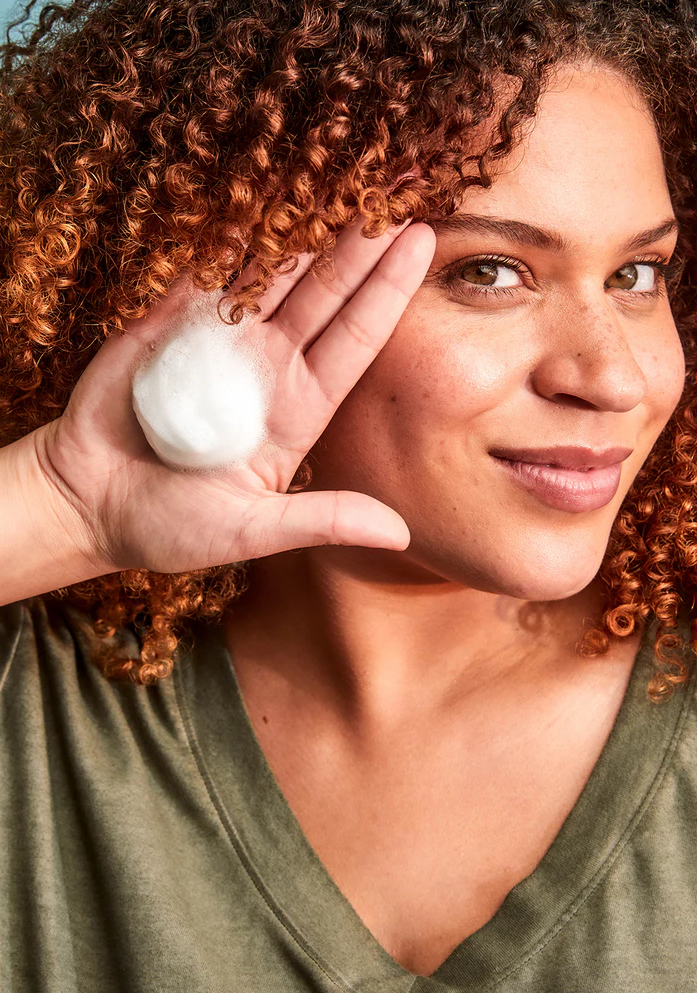 A person with curly hair holds a handful of foam or cleanser near their face, smiling at the camera.