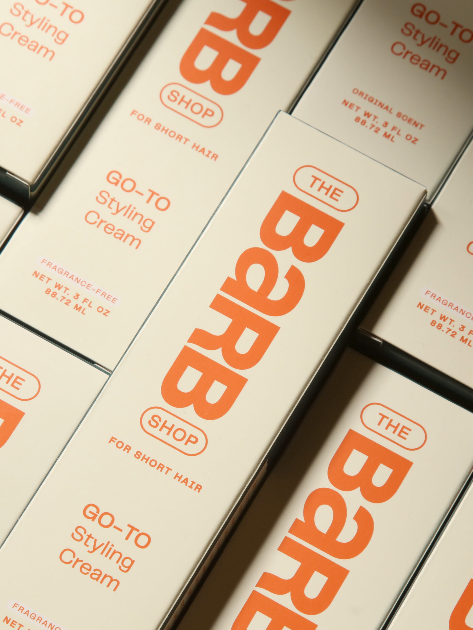 Boxes of "The Barb Shop Go-To Styling Cream" are arranged in a grid pattern, displaying product names and details in orange and light beige colors.