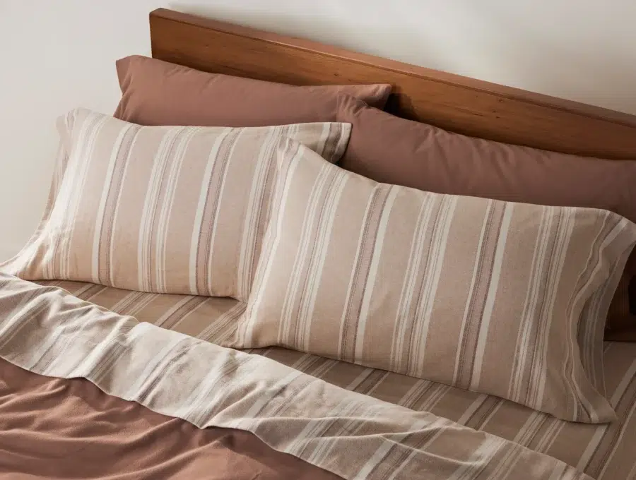 A made bed with a wooden headboard, two striped pillows, a striped sheet, and solid brown pillowcases and fitted sheet.