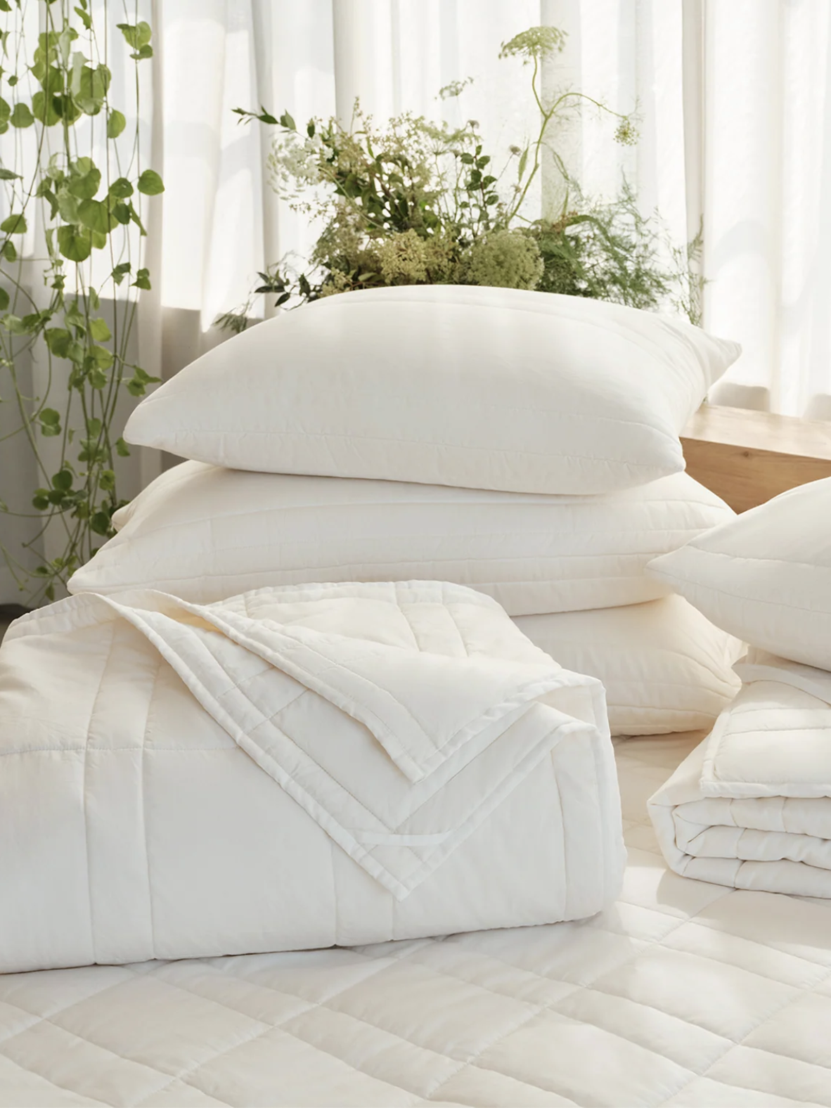 A neatly arranged bed with white comforters, folded blankets, and pillows in a sunlit room with green plants and sheer curtains in the background.