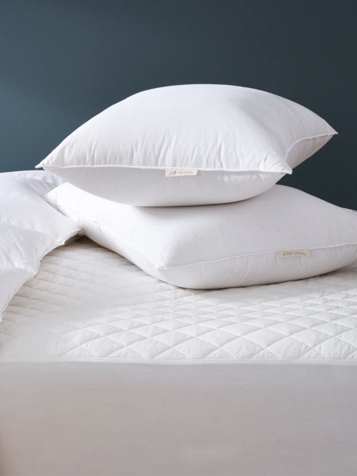 Two white pillows stacked on a neatly made bed with a white quilted mattress cover and a white comforter, against a dark blue wall.