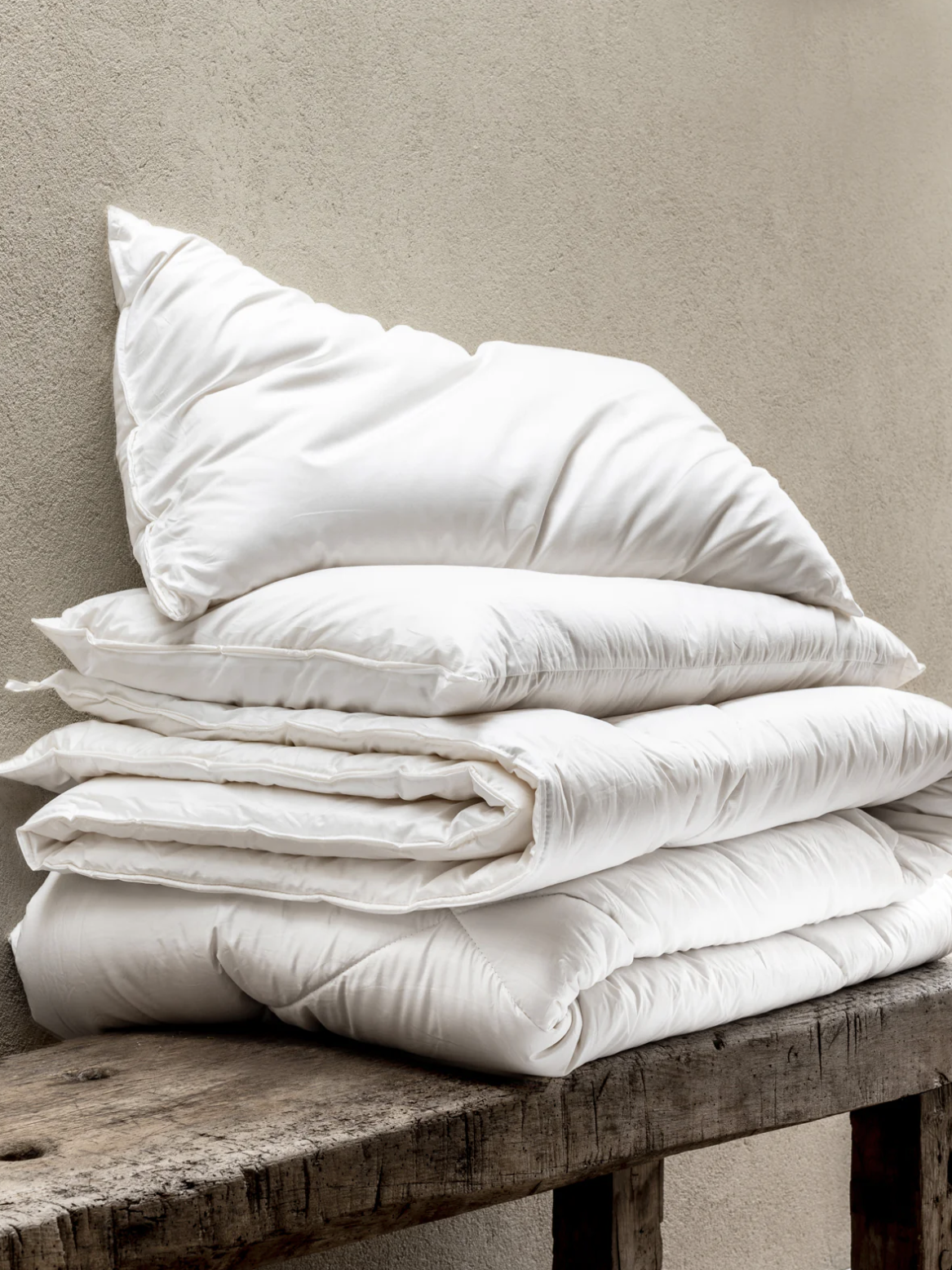 A stack of white comforters and a pillow are neatly arranged on a rustic wooden bench against a beige wall.