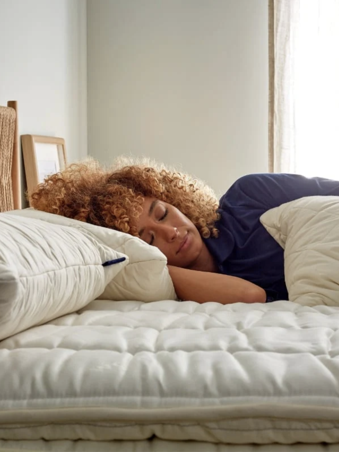 Person with curly hair sleeping on a white mattress and pillow in a softly lit bedroom.
