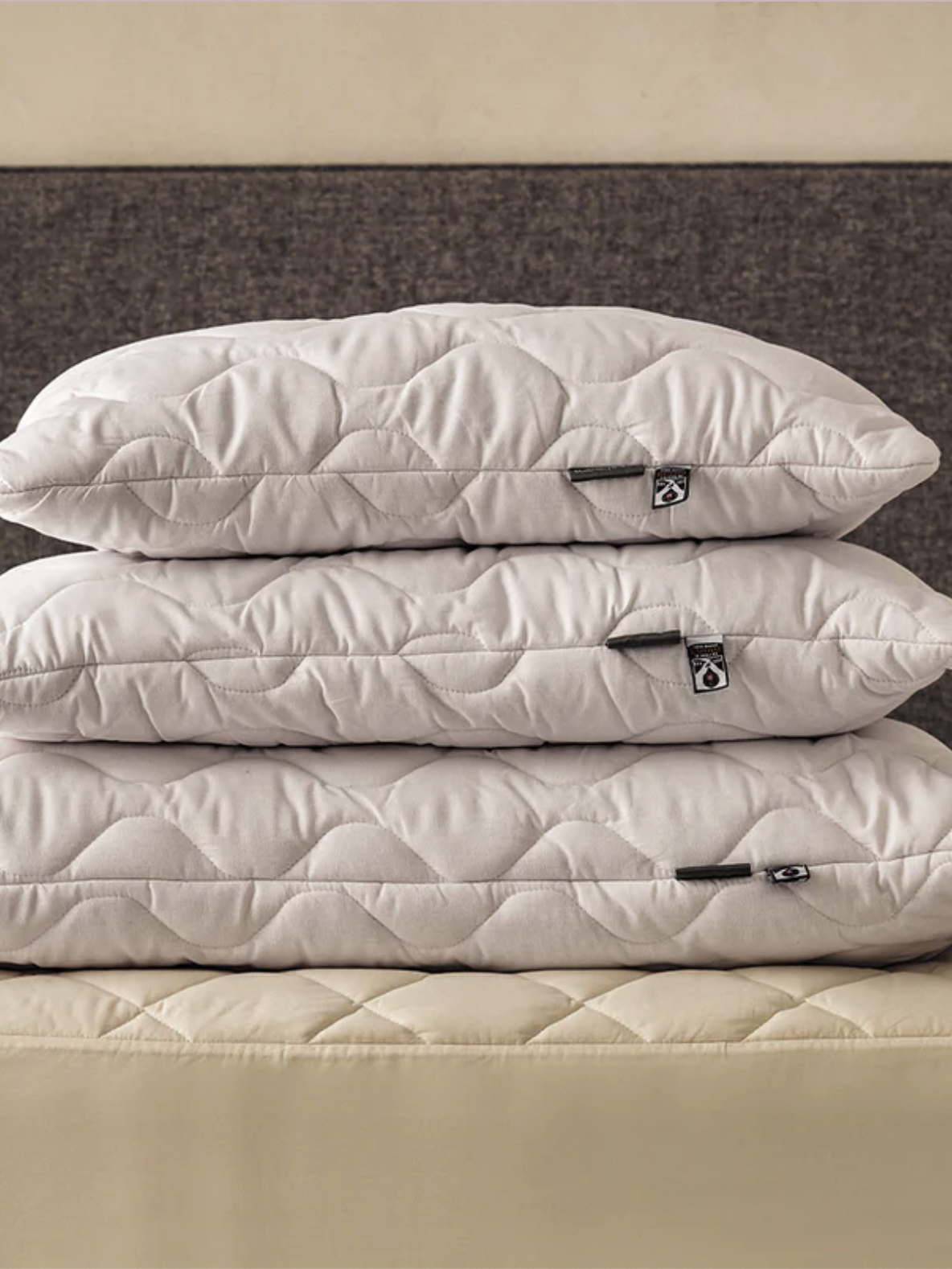 Three white quilted pillows are stacked on a beige quilted bed, with a dark gray upholstered headboard in the background.