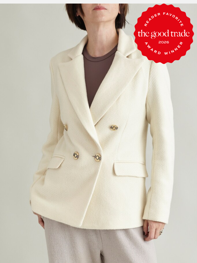 A person wearing a cream double-breasted blazer over a brown top and beige pants; a red “The Good Trade” award badge is in the top right corner.