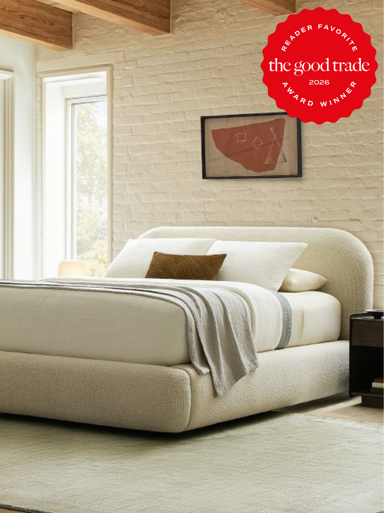 Modern bedroom with a plush upholstered bed, neutral bedding, a small brown accent pillow, and abstract wall art; “The Good Trade Reader Favorite 2026 Award Winner” badge in the corner.