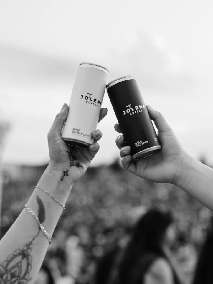 Two hands holding up Jolene canned drinks in a toast at an outdoor event, with a blurred crowd in the background.