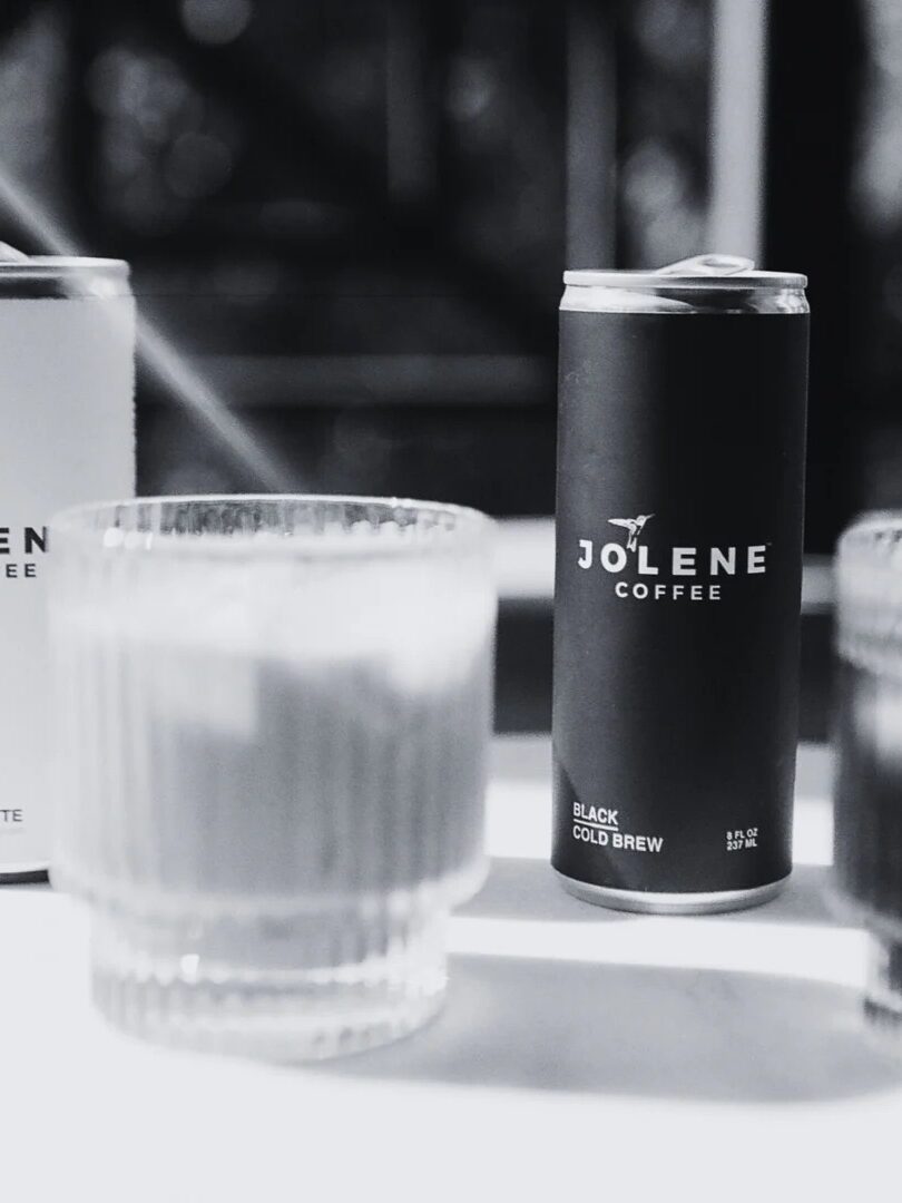 Two cans of Jolene Coffee, one labeled “Latte” and the other “Black Cold Brew,” are placed on a table beside two glass tumblers. The image is in black and white.