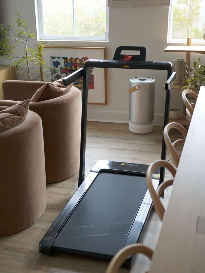 A treadmill is placed in a living room with light wood flooring, near two brown chairs and a window with natural light coming in.