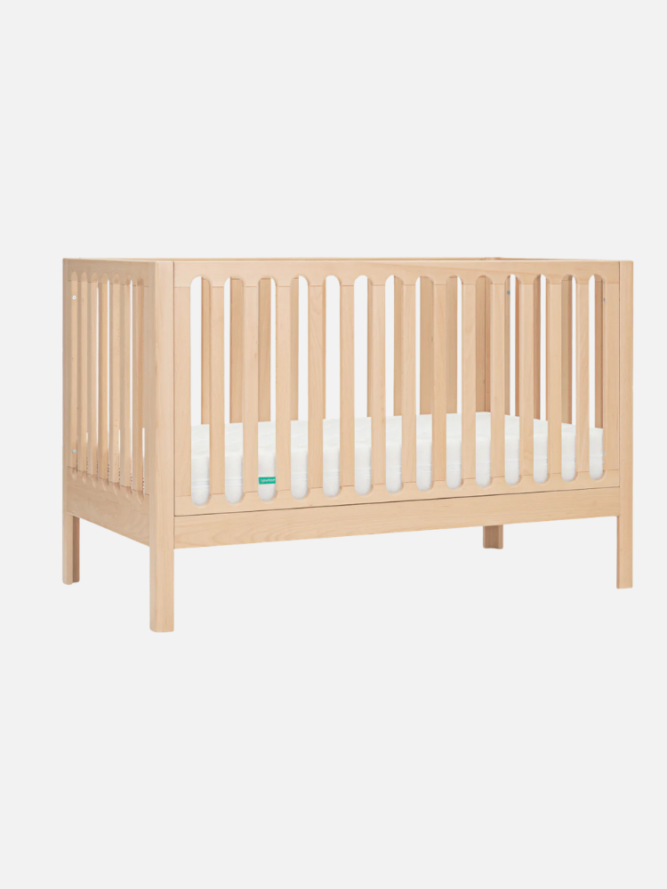 A wooden baby crib with vertical slats on all sides and a white mattress, set against a plain white background.
