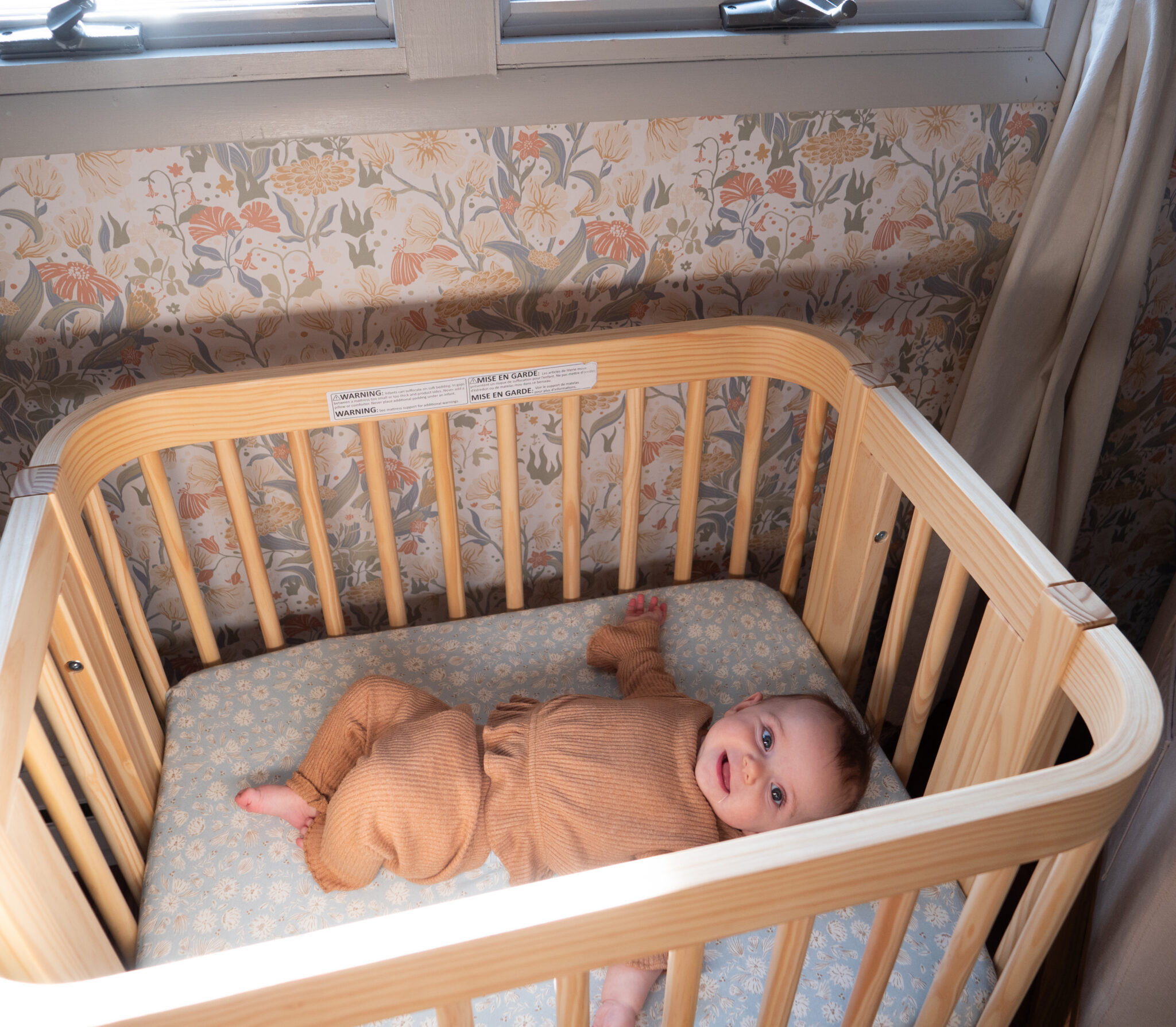 A baby in brown clothing lies on its back in a light wooden crib, with floral wallpaper and a window in the background.