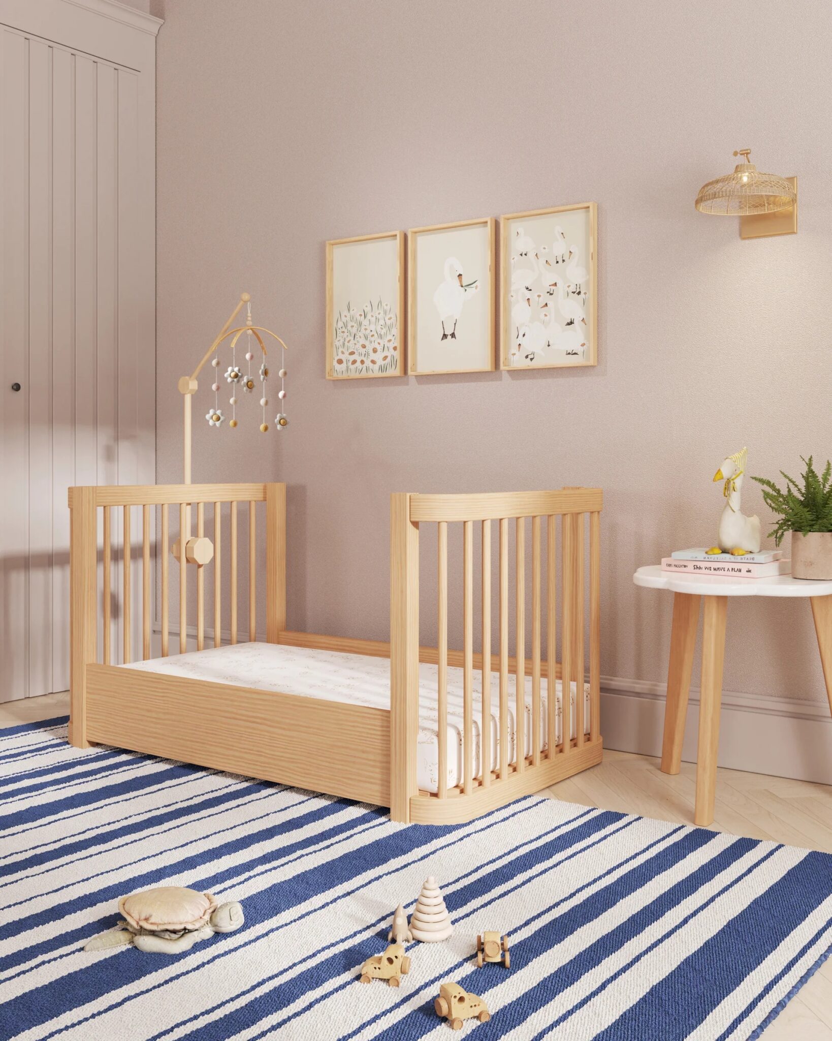 A wooden toddler bed with a mobile stands on a blue and white striped rug in a nursery, with toys on the floor and framed art on the wall.