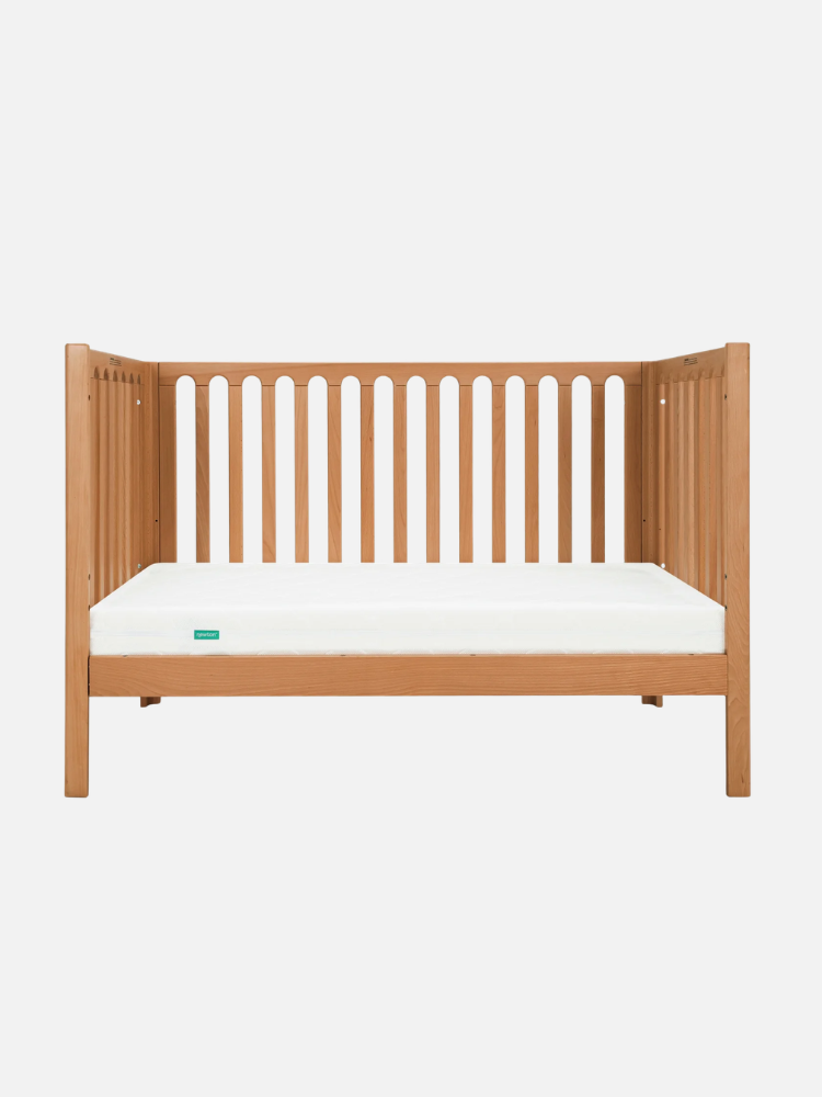 A wooden crib with a light natural finish, open slats along the back and sides, and a plain white mattress on a white background.