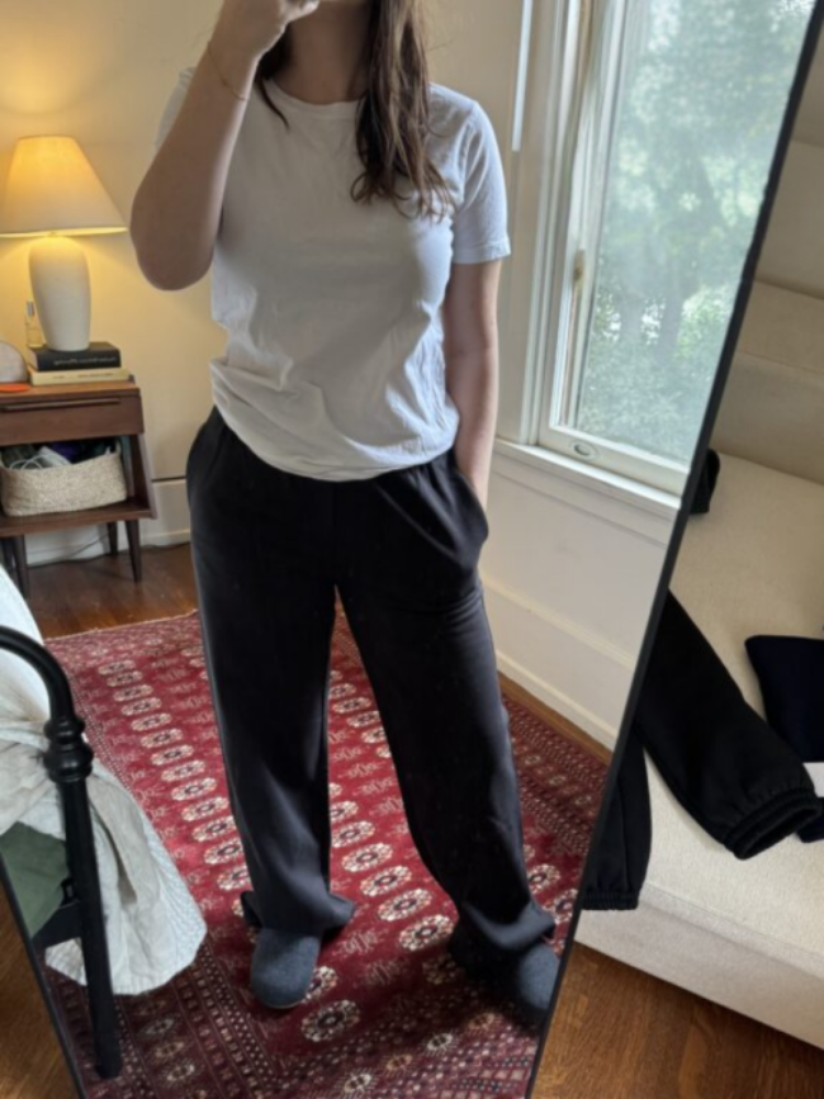 Person stands in front of a mirror wearing a white T-shirt, black pants, and dark slippers in a bedroom with a red patterned rug and a window in the background.