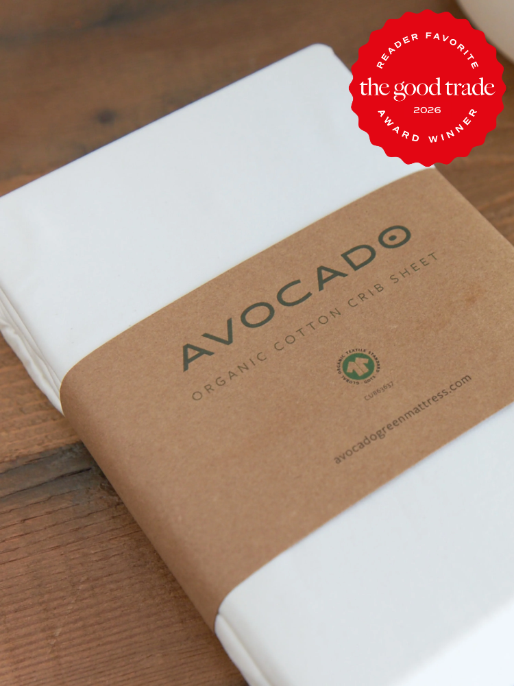 A packaged Avocado brand organic cotton crib sheet with a brown paper label, placed on a wooden surface. A red “the good trade award winner 2026” badge is in the top right corner.