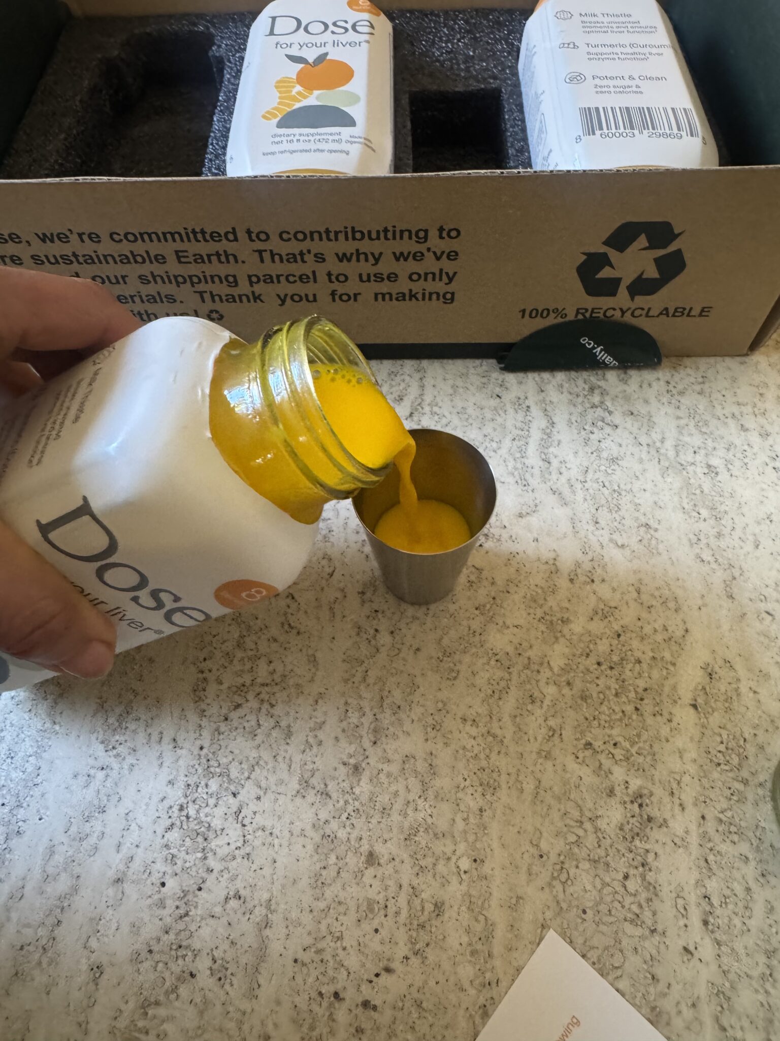 A hand pours yellow liquid from a Dose bottle into a metal measuring cup, with Dose packaging and a recyclable box in the background.