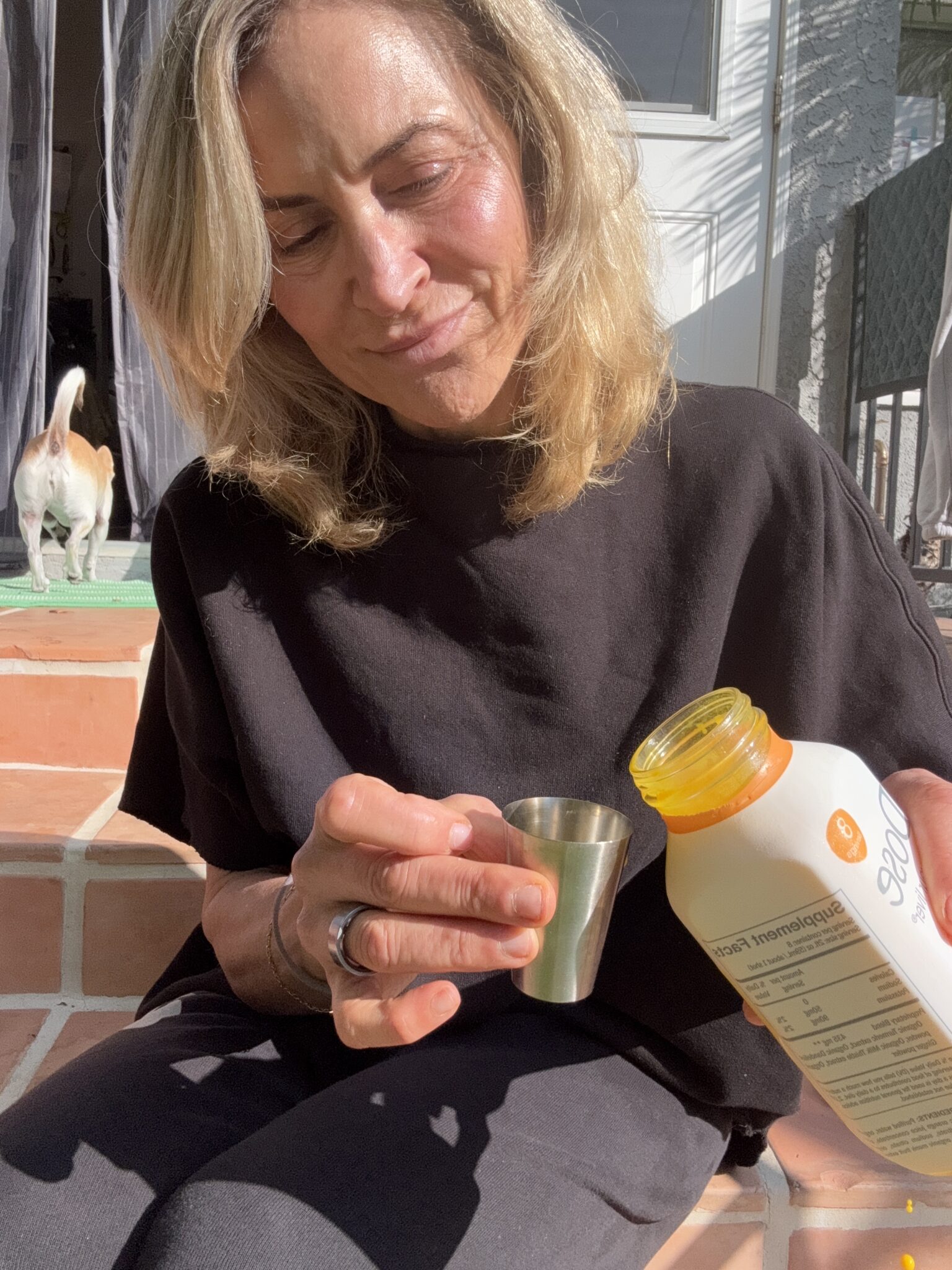 A woman sitting on outdoor steps pours a yellow liquid from a bottle into a metal measuring cup; a dog stands in the background.