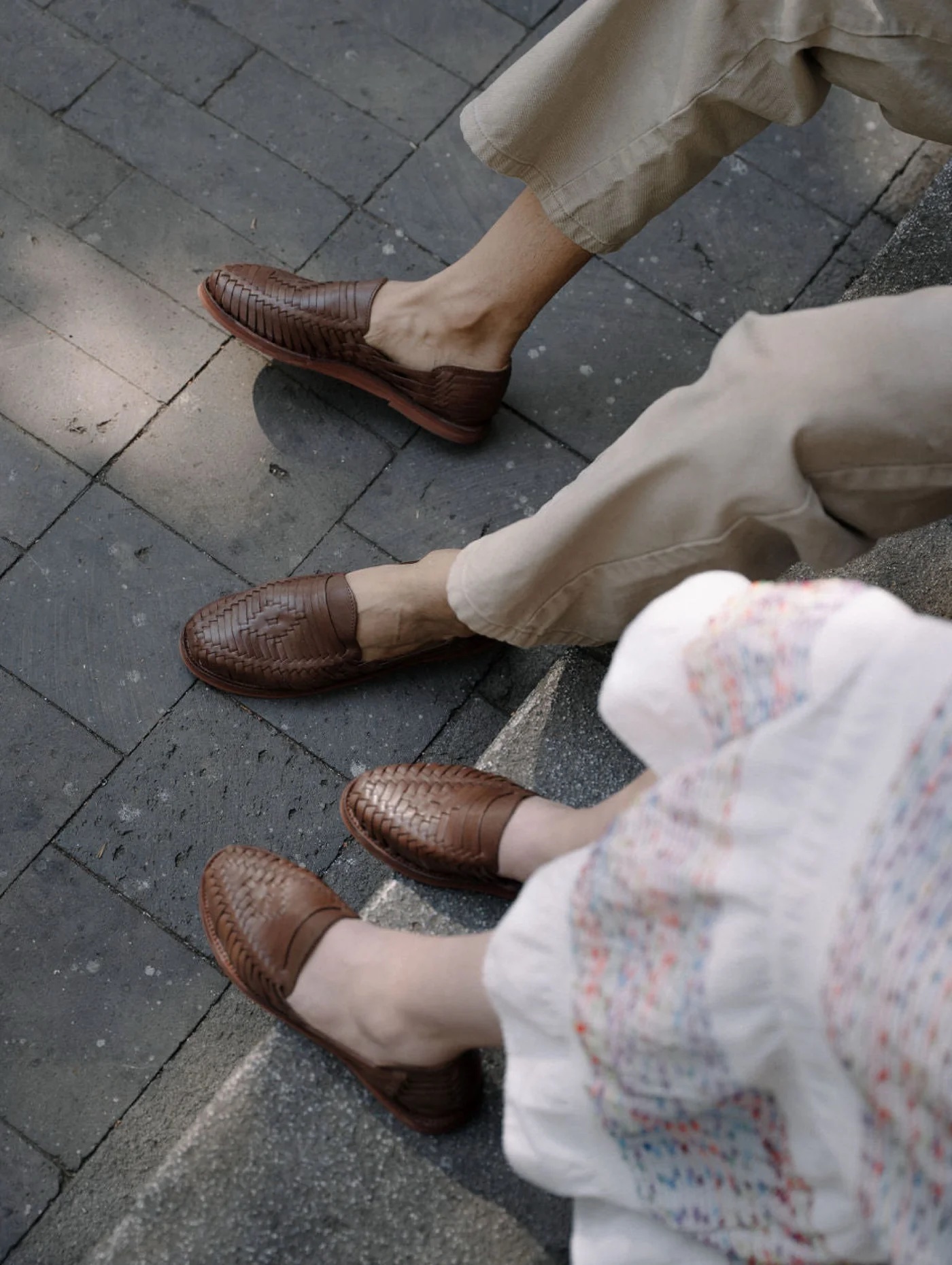 Two people showcase brown woven leather huarache sandals; one pairs them with light pants, the other with a light, patterned dress, as they sit together on steps against a stone pavement backdrop.