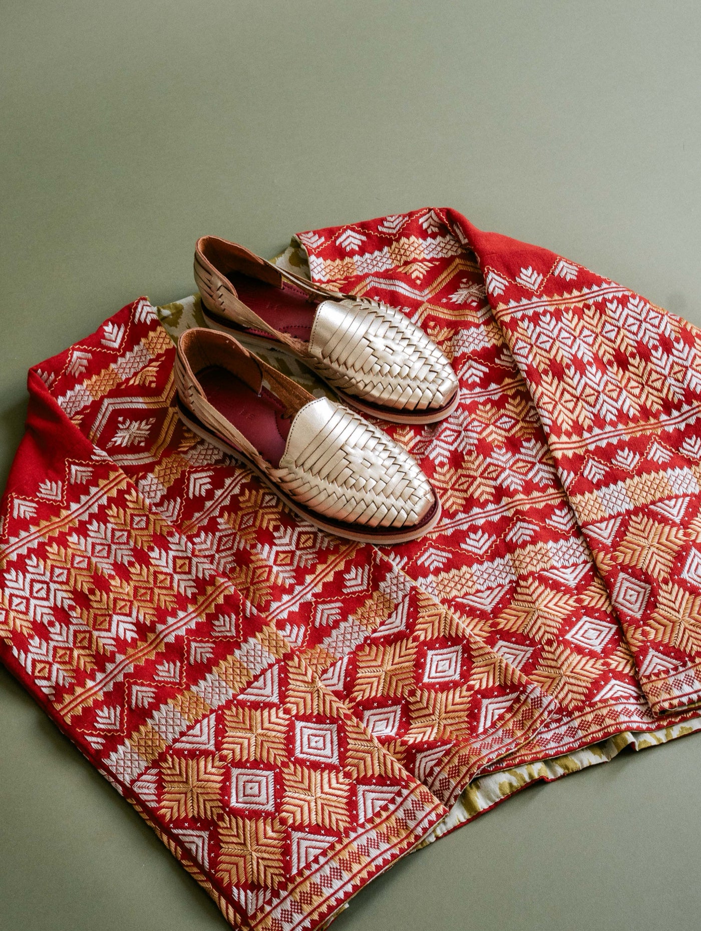 A pair of woven leather huarache sandals is placed on top of a red and gold patterned textile, both resting on a green surface.