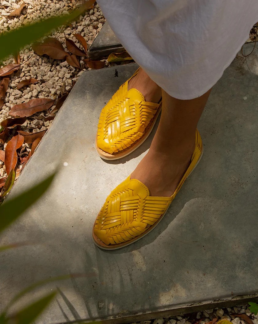 Person wearing a white skirt and bright yellow woven leather huarache sandals standing on a stone path with pebbles and leaves around.