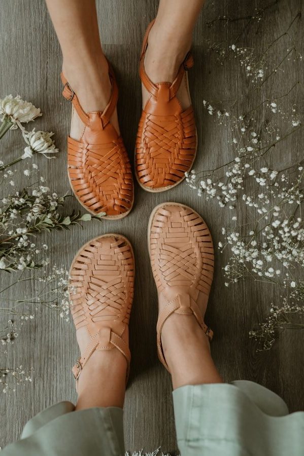 Two people wearing woven huarache sandals stand on a wooden floor, surrounded by small white flowers and greenery scattered around their feet.