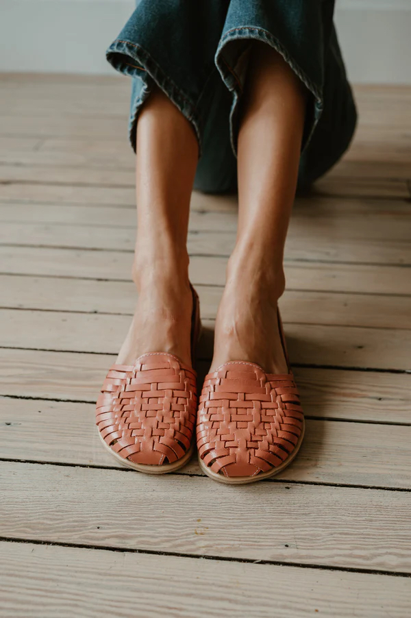 Person wearing woven, tan leather huarache sandals and cropped blue jeans, seated on a light wooden floor.