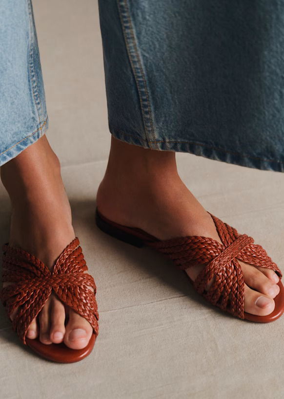 Person wearing blue denim jeans and brown woven leather huarache sandals, standing on a light-colored floor.