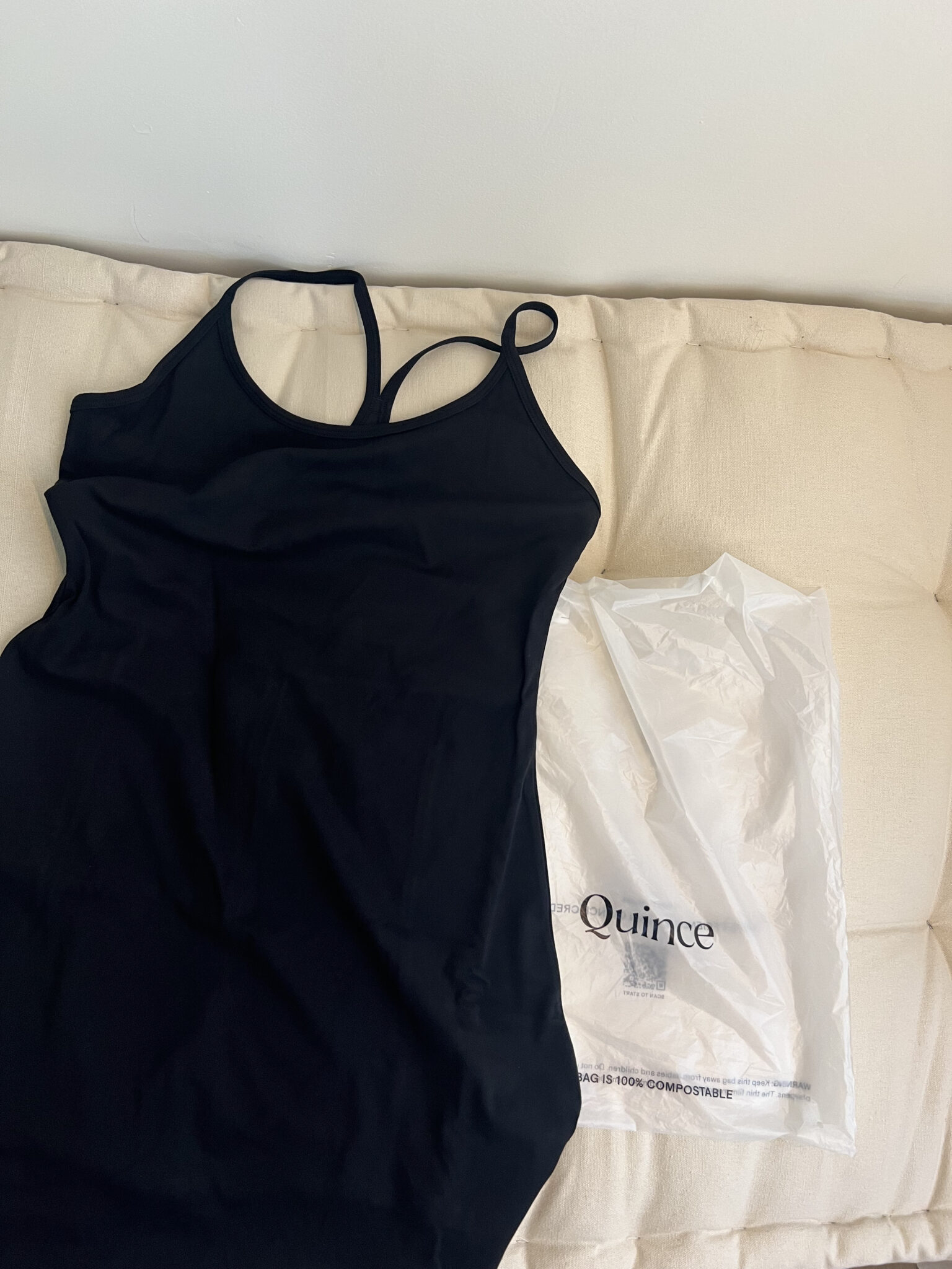 A black sleeveless dress with thin straps lies next to a white compostable plastic bag labeled "Quince" on a beige cushioned surface.