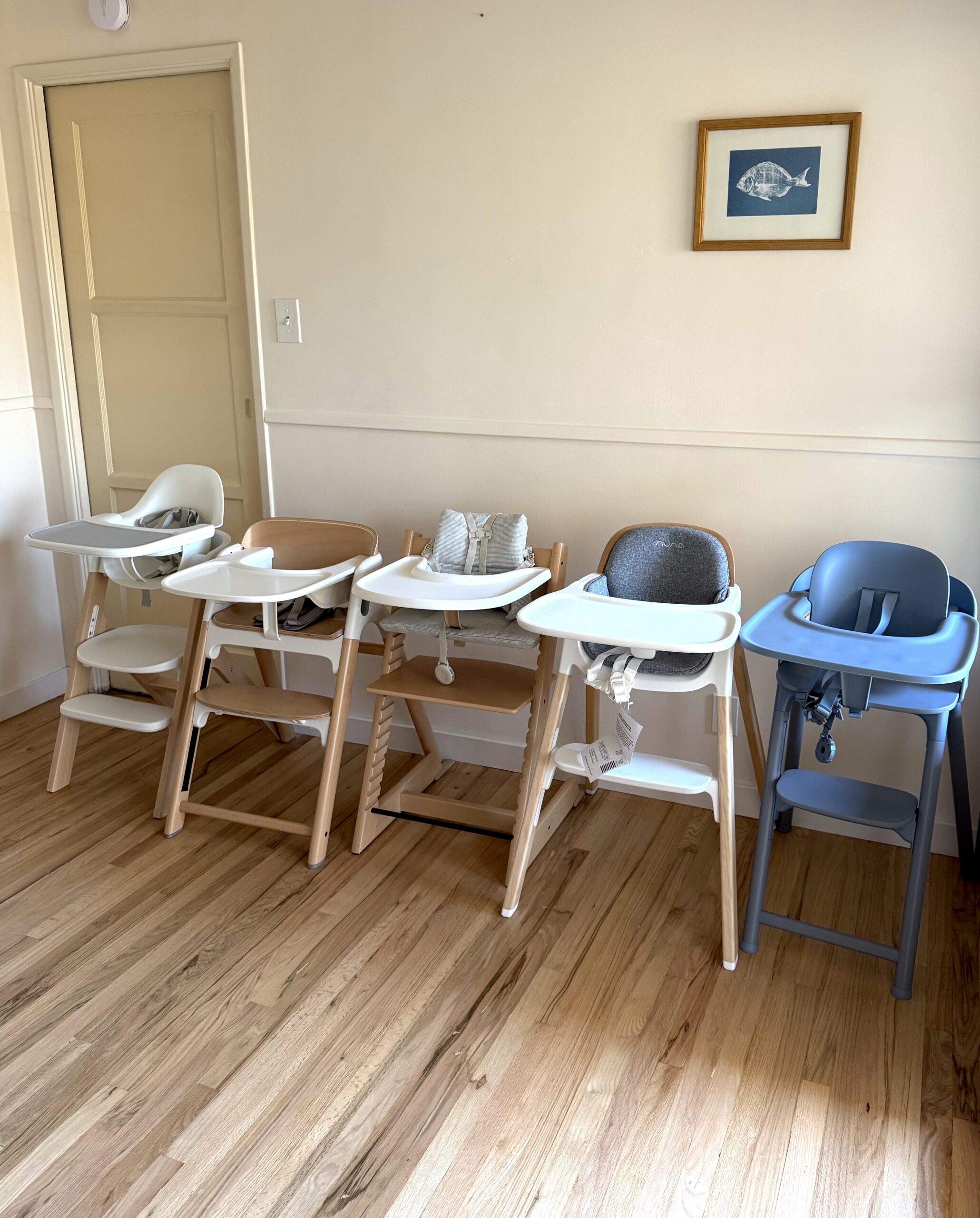 Four high chairs in different styles and colors are lined up against a wall on a wooden floor, with a framed fish print hanging above.