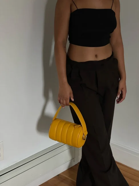 Person standing indoors, wearing a black crop top and dark pants, holding a small yellow handbag by their side.