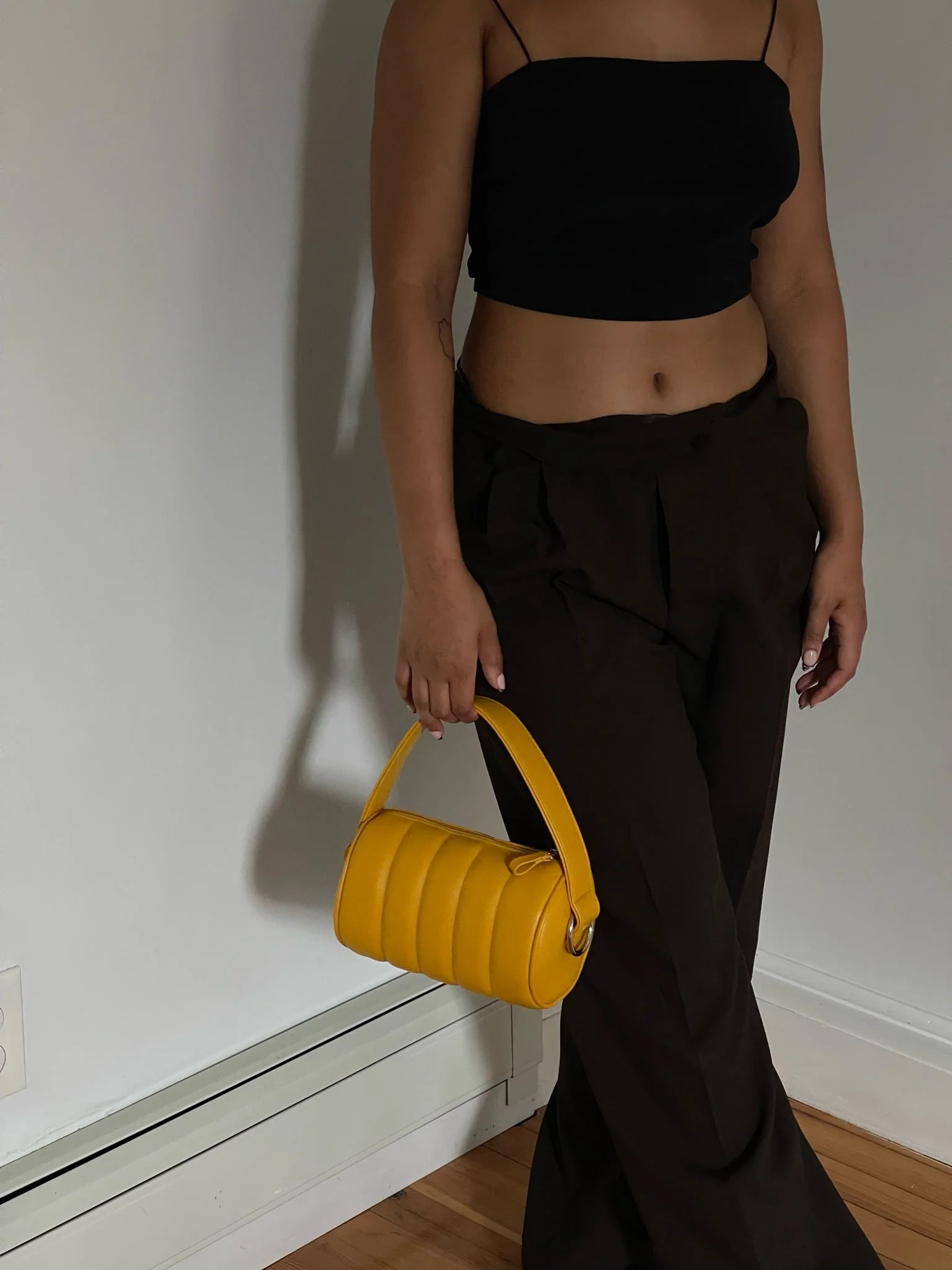 Person standing indoors, wearing a black crop top and dark pants, holding a small yellow handbag by their side.