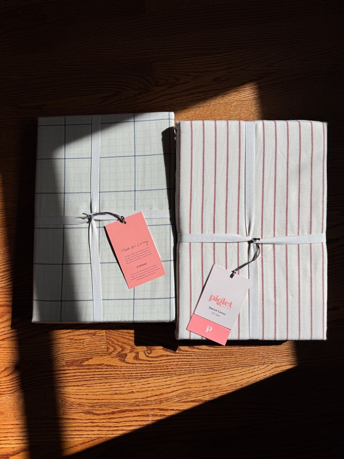 Two folded and packaged sets of bed linens with tags, one with a gray grid pattern and the other with red vertical stripes, placed on a wooden floor in natural light.