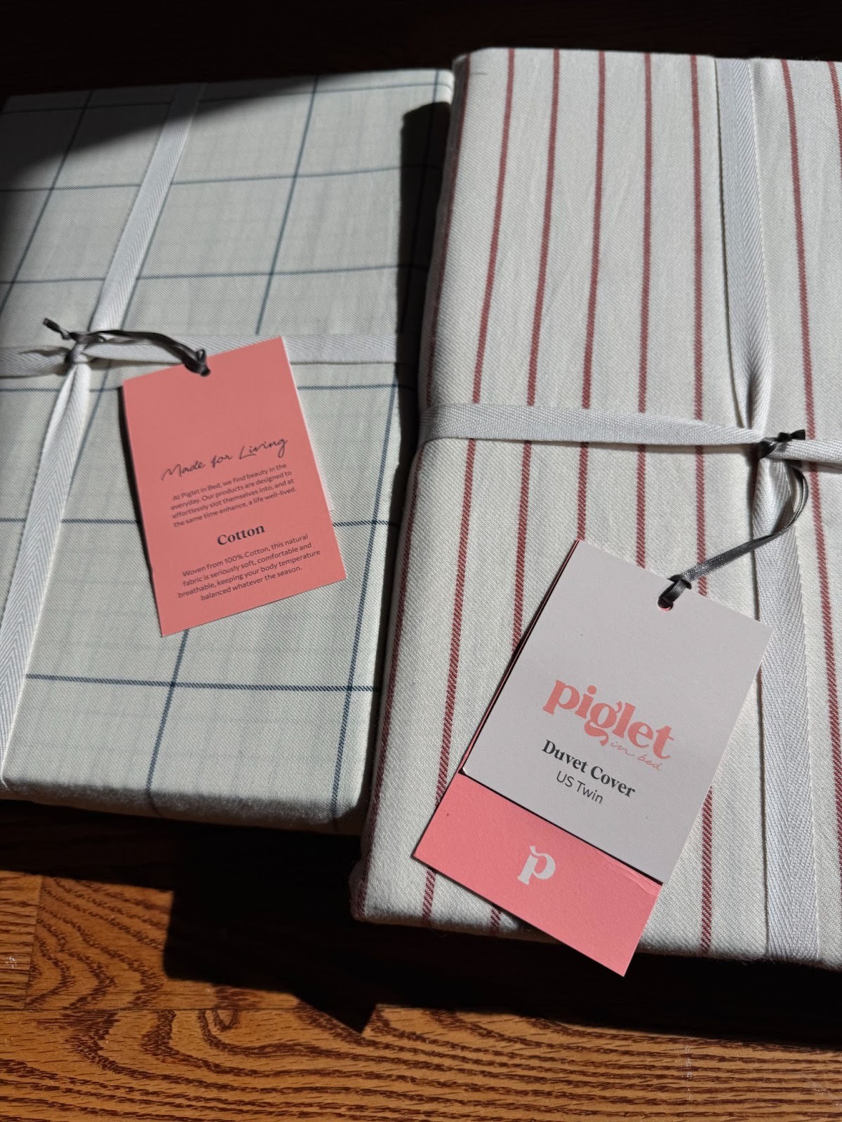 Two packaged duvet covers with pink tags; one has a blue grid pattern, the other has red stripes. Both are labeled "piglet" and are laying on a wooden surface.