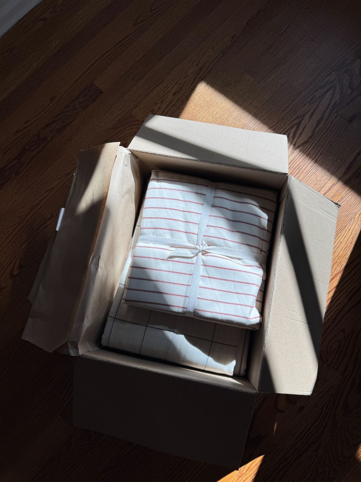 An open cardboard box on a wooden floor contains folded, ribbon-tied fabric with red stripes, partially lit by sunlight.