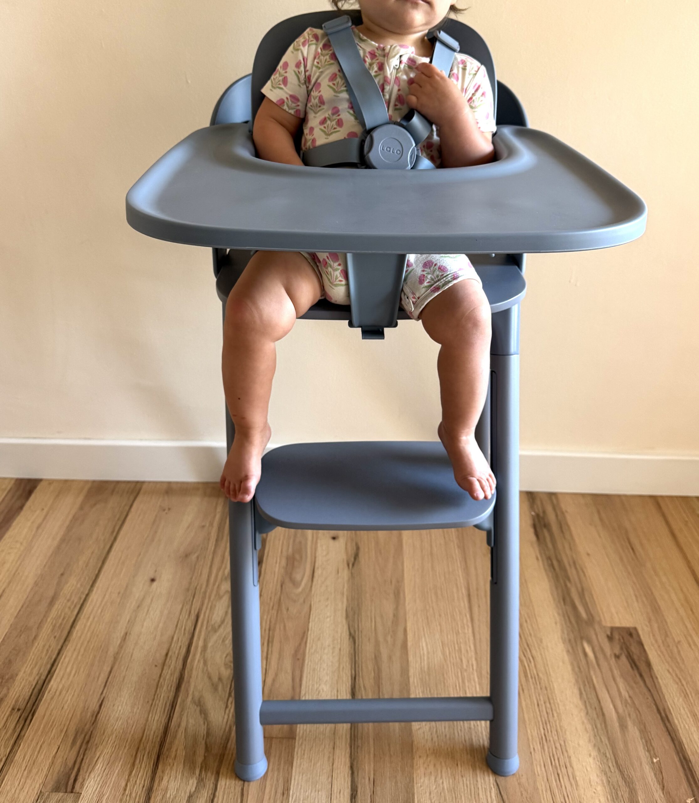 A baby wearing a floral onesie sits in a gray high chair with a safety harness, feet resting on the footrest, against a light wall and wooden floor.
