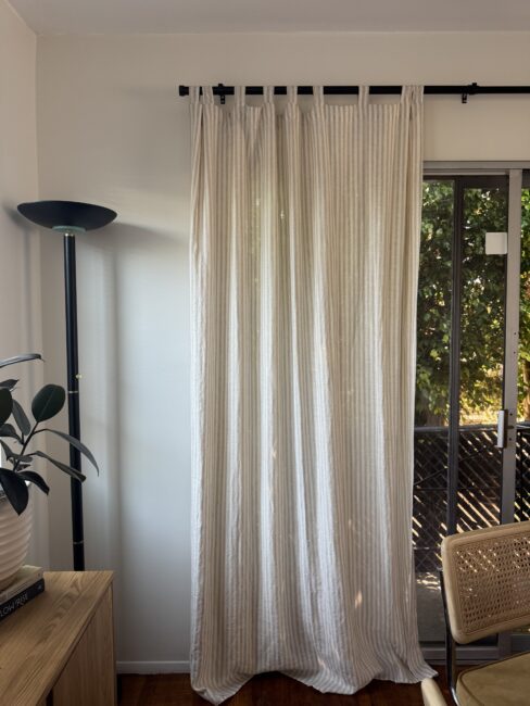 A beige striped curtain hangs in front of a window with a sliding glass door; a floor lamp, plant, and wooden furniture are also visible in the room.