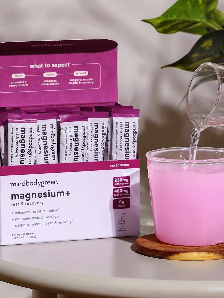 A hand pours water into a cup containing a pink powder drink next to an open box of mindbodygreen magnesium+ supplement packets on a table.