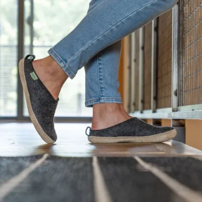 Person wearing gray slip-on shoes and light blue jeans, standing indoors on a wooden floor next to a wall with vertical metal bars.