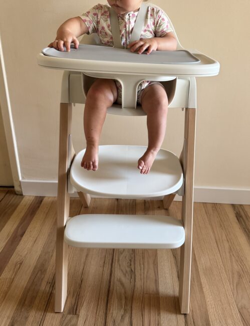 A baby sits in a modern high chair with a tray, wearing a patterned outfit. Only the baby’s torso, arms, and legs are visible. The chair is on a wooden floor against a plain wall.
