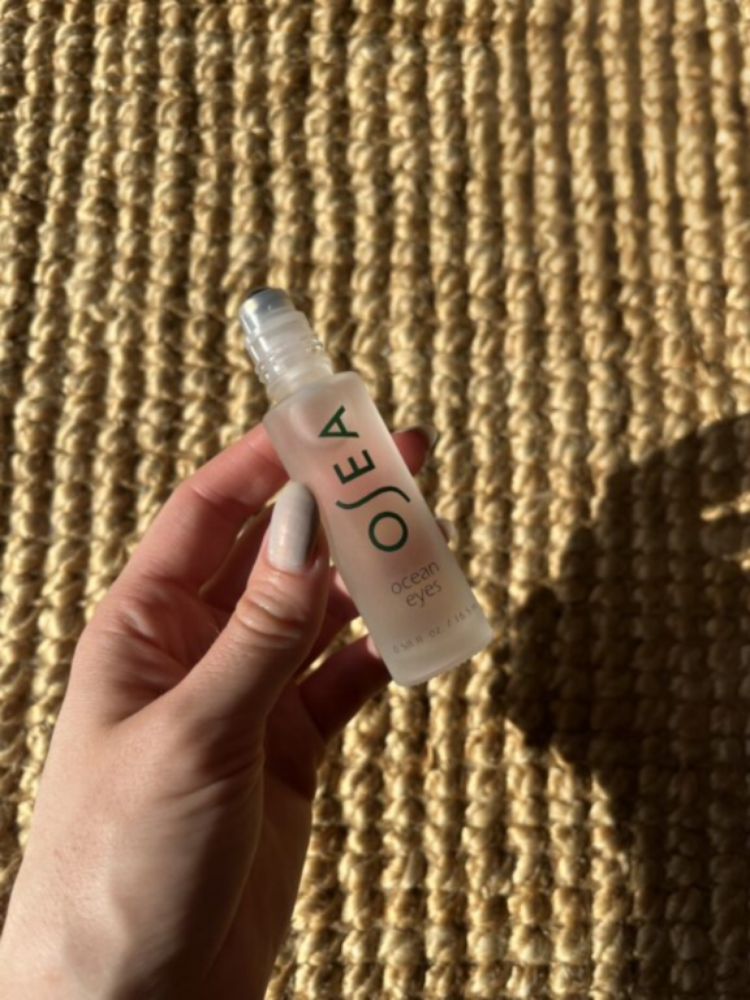 A hand holding a clear OSEA Ocean Eyes serum bottle with a rollerball top, against a woven textured surface in sunlight.