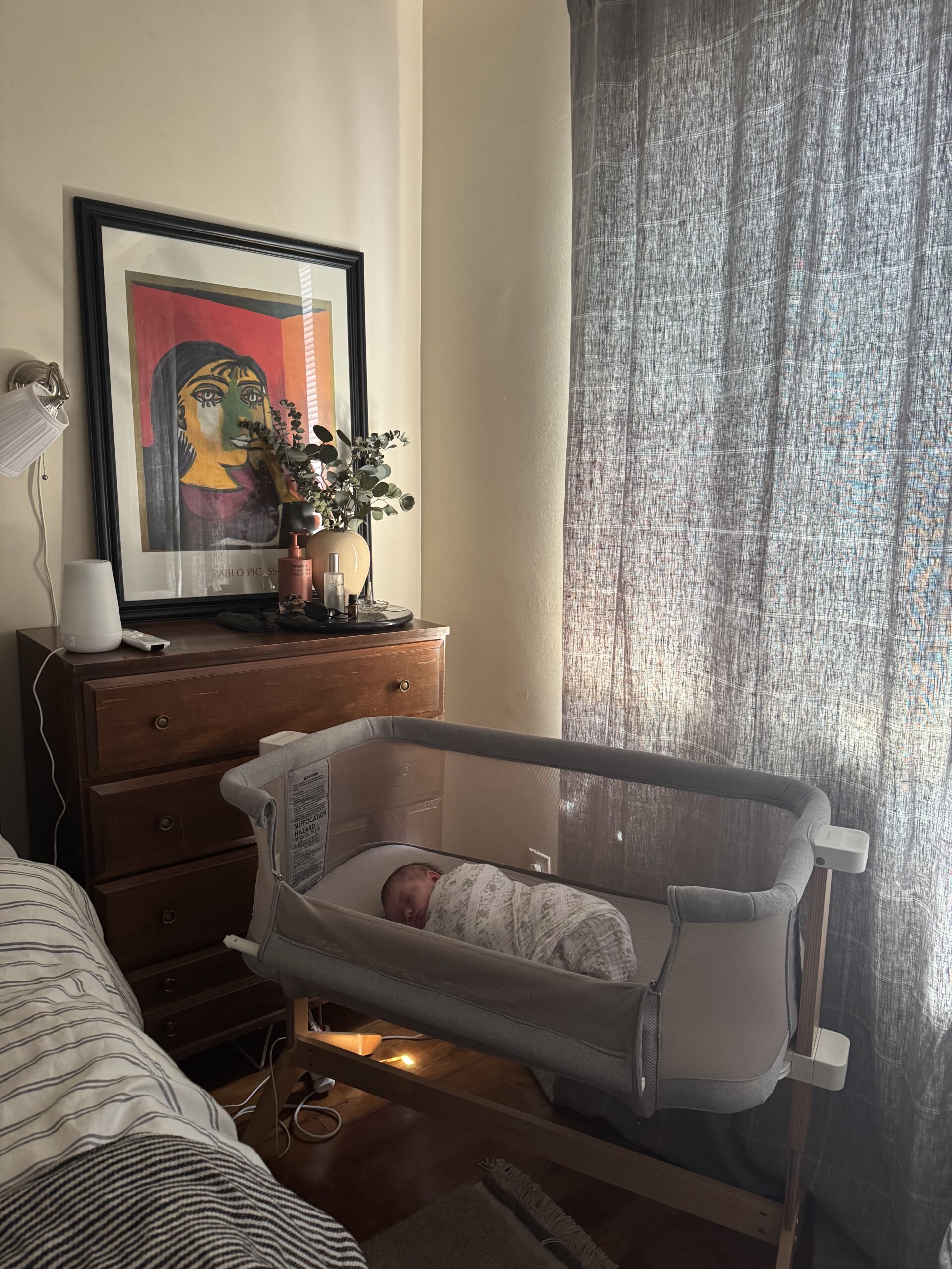 A swaddled baby lies in a gray bassinet next to a bed, with sunlight filtering through curtains and a dresser topped with decor and framed art in the background.