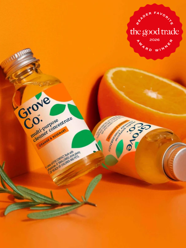 Two bottles of Grove Co. multi-purpose cleaner concentrate with orange and rosemary scents are displayed beside an orange half and rosemary sprig on an orange background.