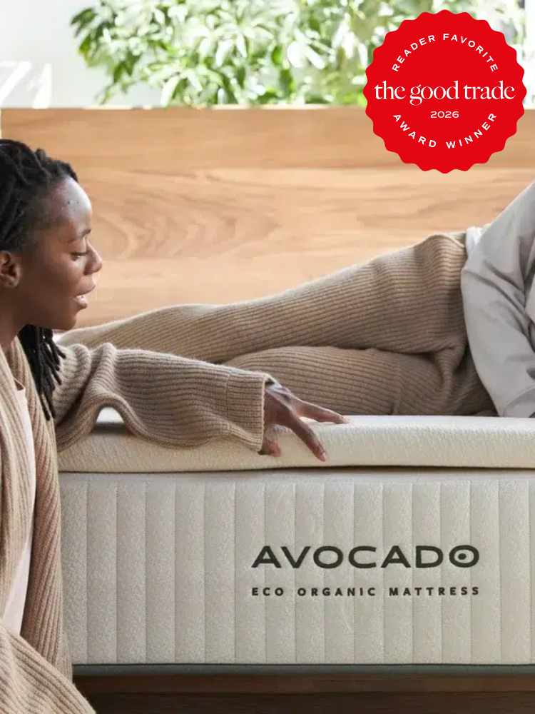 Two people in loungewear sit on and beside an Avocado Eco Organic Mattress in a modern bedroom. A "The Good Trade 2026 Reader Favorite" award badge is shown in the top right corner.