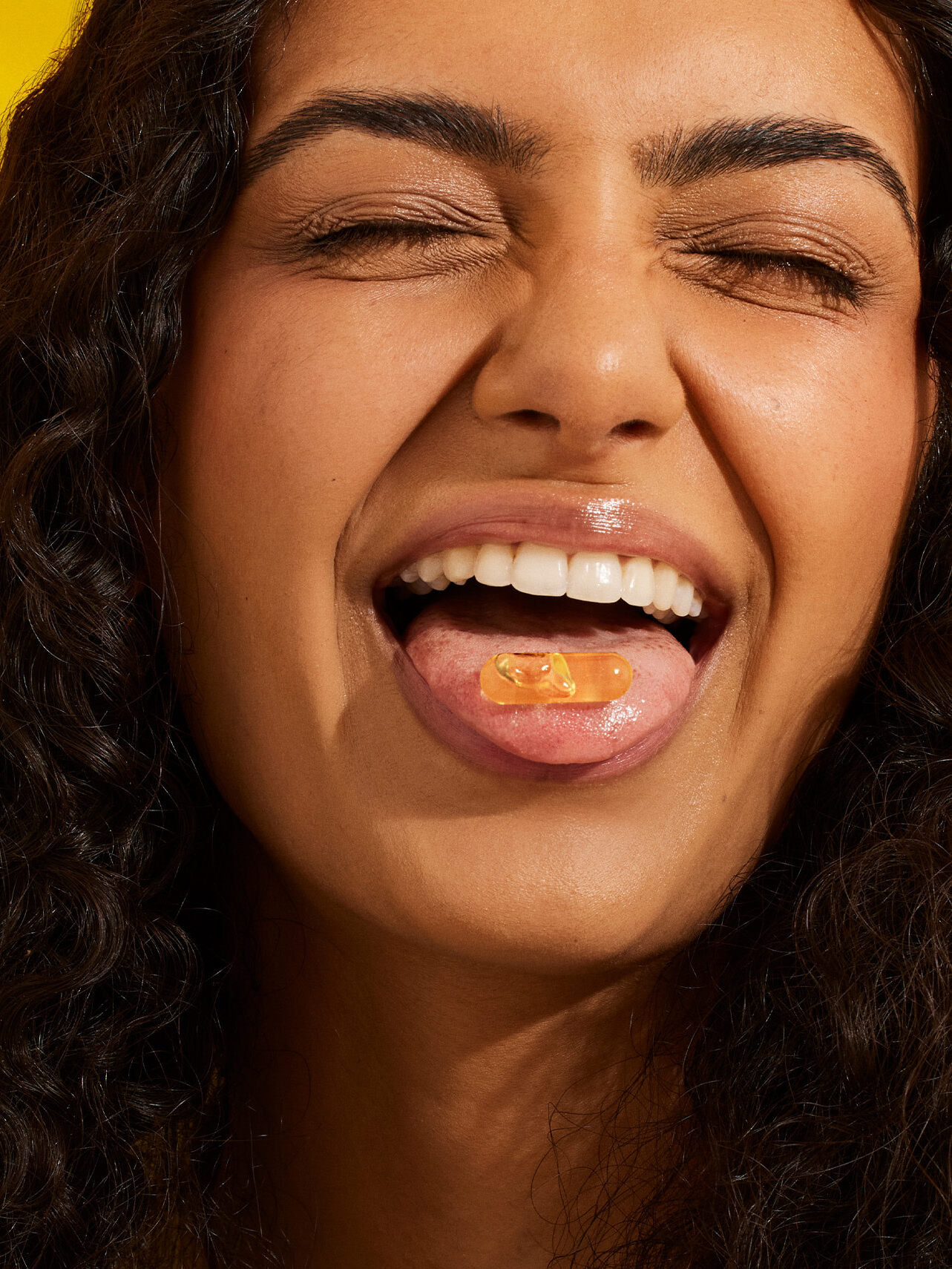 A person with curly hair smiles with eyes closed, sticking out their tongue to show a yellow capsule resting on it, against a yellow background.