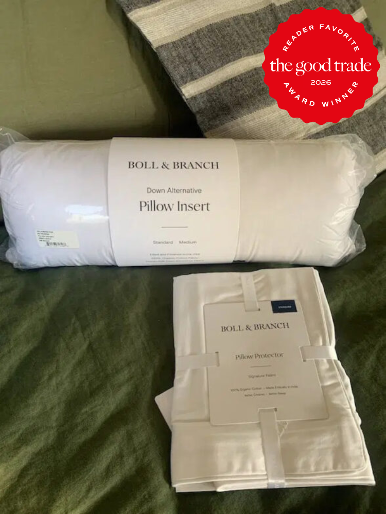A Boll & Branch down alternative pillow insert and pillow protector are displayed on a bed with green and striped bedding. A 2026 "The Good Trade Reader Favorite" badge is in the top right corner.