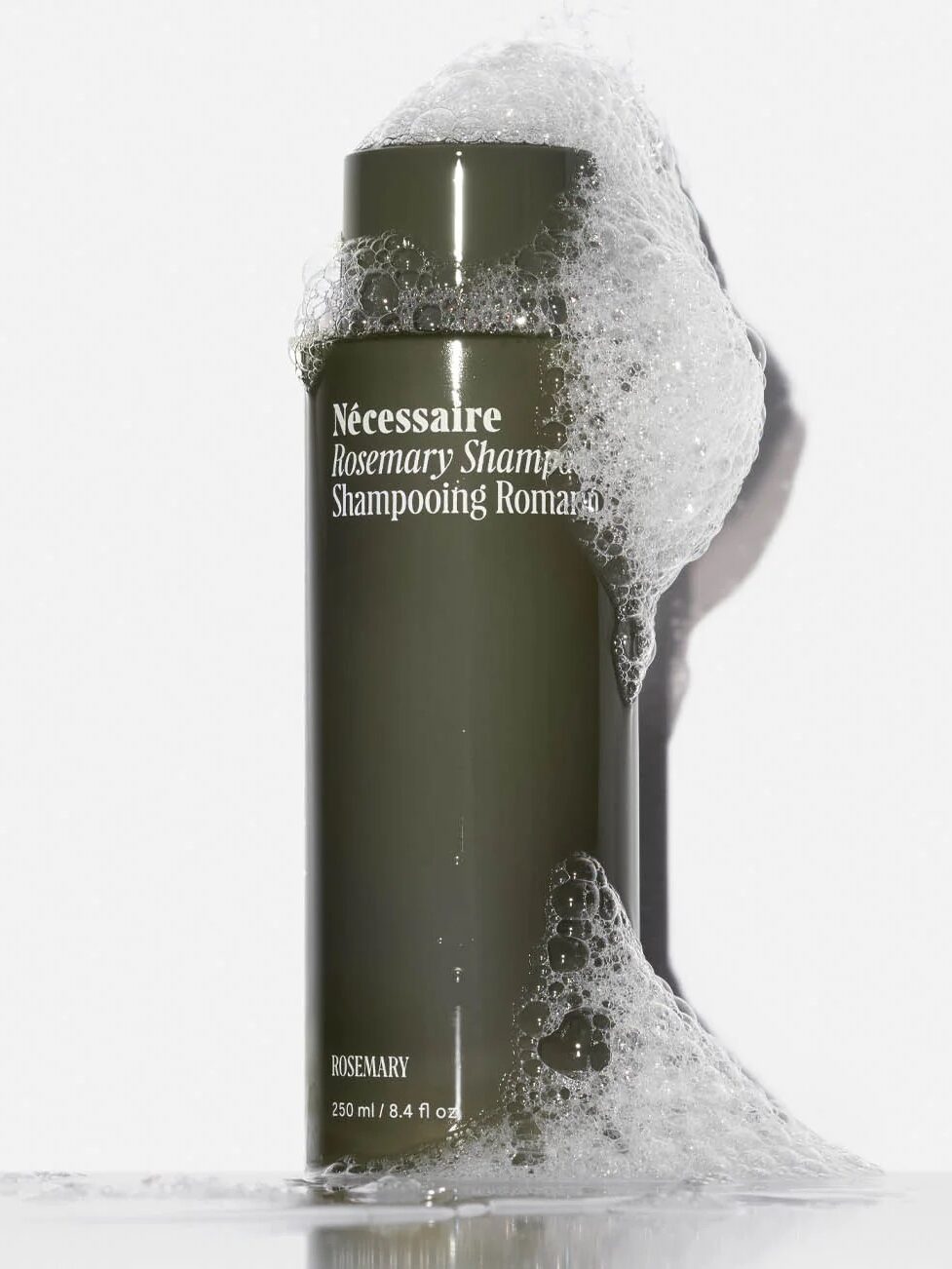 A green bottle of Nécessaire Rosemary Shampoo is covered in soap suds against a plain white background.