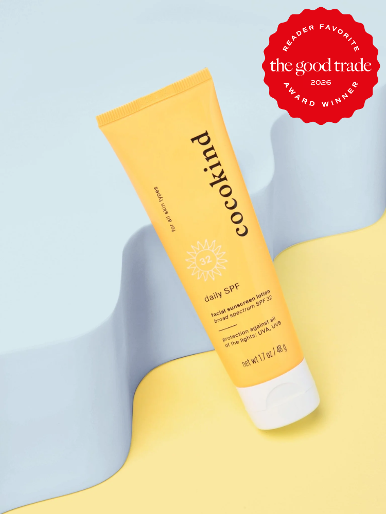 A yellow tube of Cocokind daily SPF 32 sunscreen lotion is displayed on a wavy pastel blue and yellow background with a "The Good Trade Award Winner 2026" badge in the corner.
