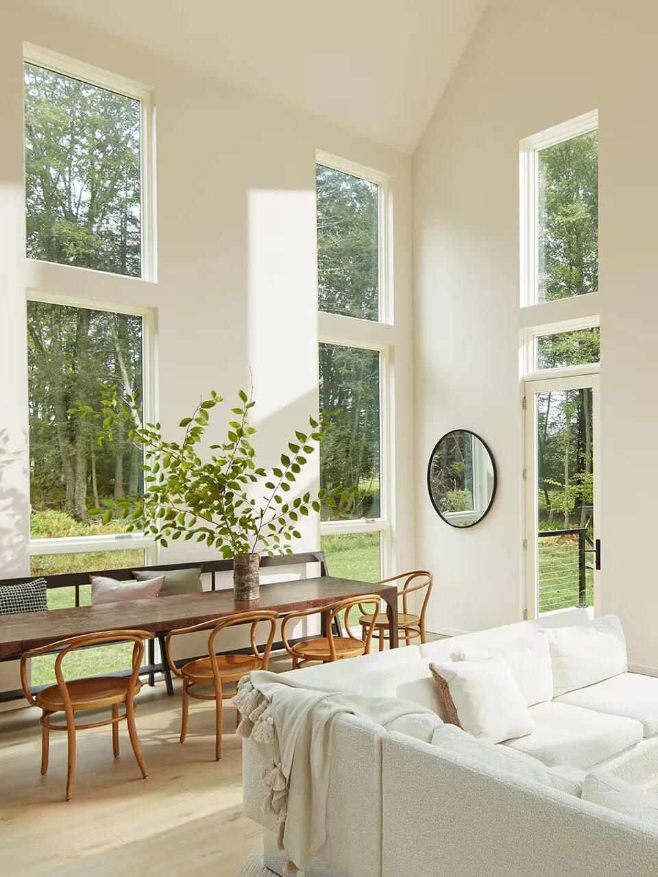 Bright, modern living and dining area with large windows, a white sofa, wooden dining table with chairs, and a tall vase with green branches.